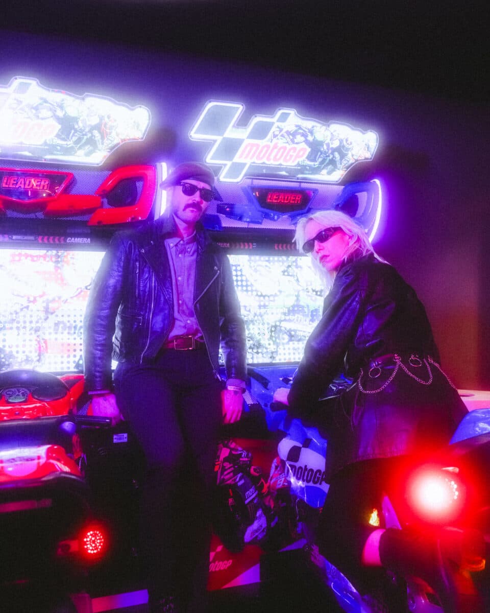 Two people in leather jackets pose beside neon-lit MotoGP arcade games in a dimly lit setting.