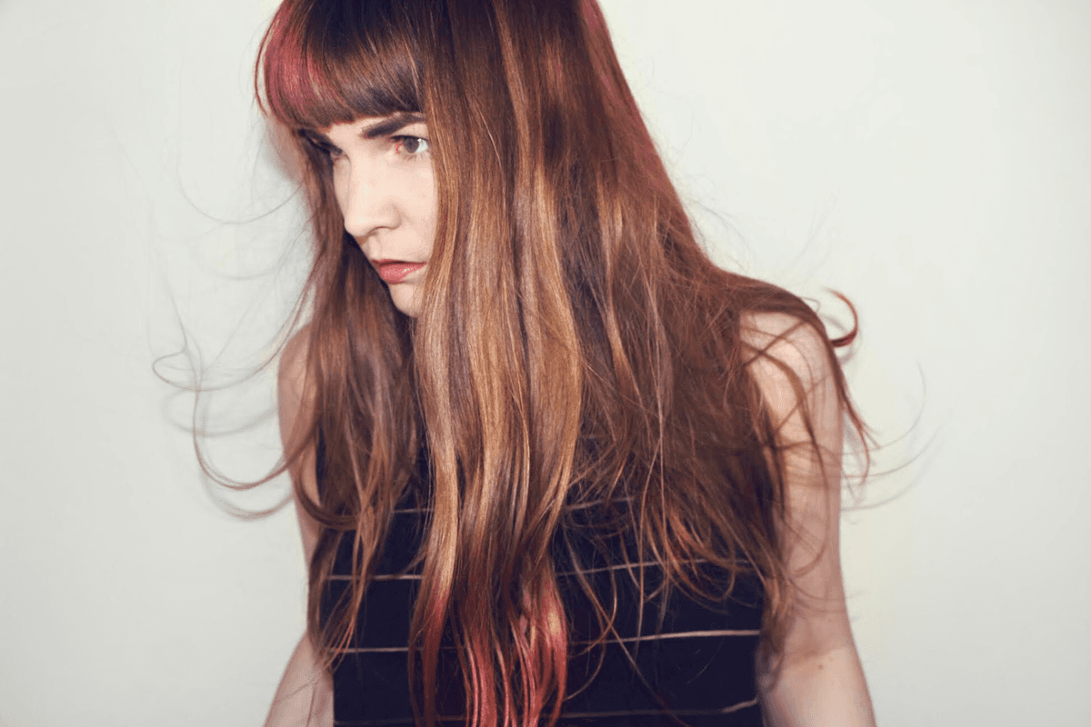 A person with long, reddish-brown hair and bangs is looking to the side against a plain background.