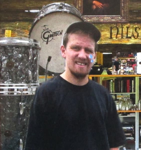 A person wearing a black shirt and cap stands in front of a drum set and shelves with various items.