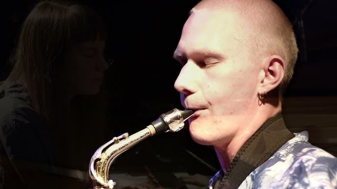 A person is playing a saxophone with eyes closed, and a faint silhouette of another person is in the background.