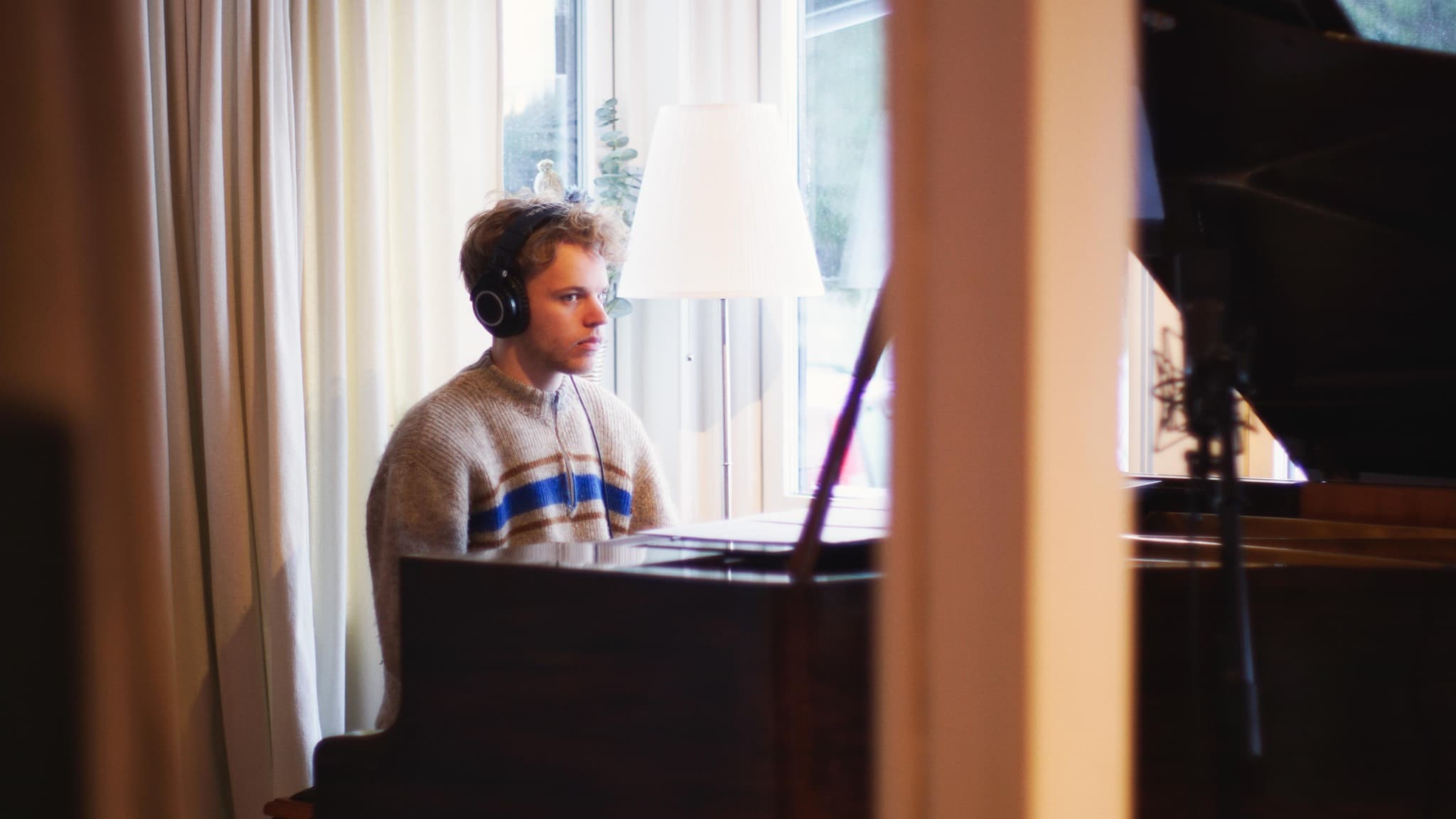 A person wearing headphones and a sweater sits at a piano near a window with a floor lamp in the background.