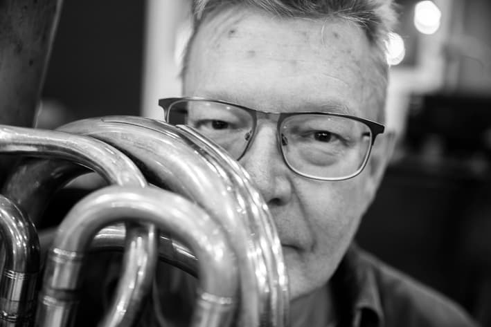 A man with glasses is partially obscured by the loops of a Tuba.