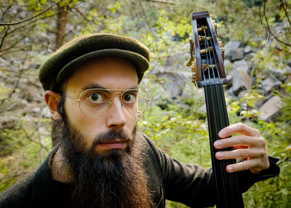 A person with glasses and a beard, wearing a cap, holds a stringed instrument in a forest setting.