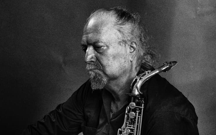 An older man with a beard holds a saxophone, looking contemplative in a black and white portrait.