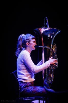 A person is seated, playing a large brass instrument, possibly a tuba, against a dark background.