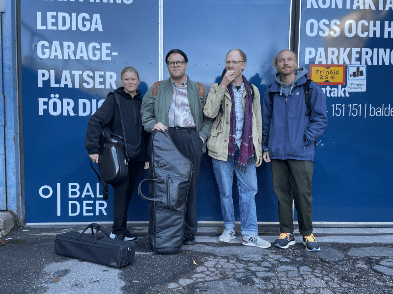 Four people stand in front of a blue sign, each holding instrument cases, with a cracked pavement in the foreground.