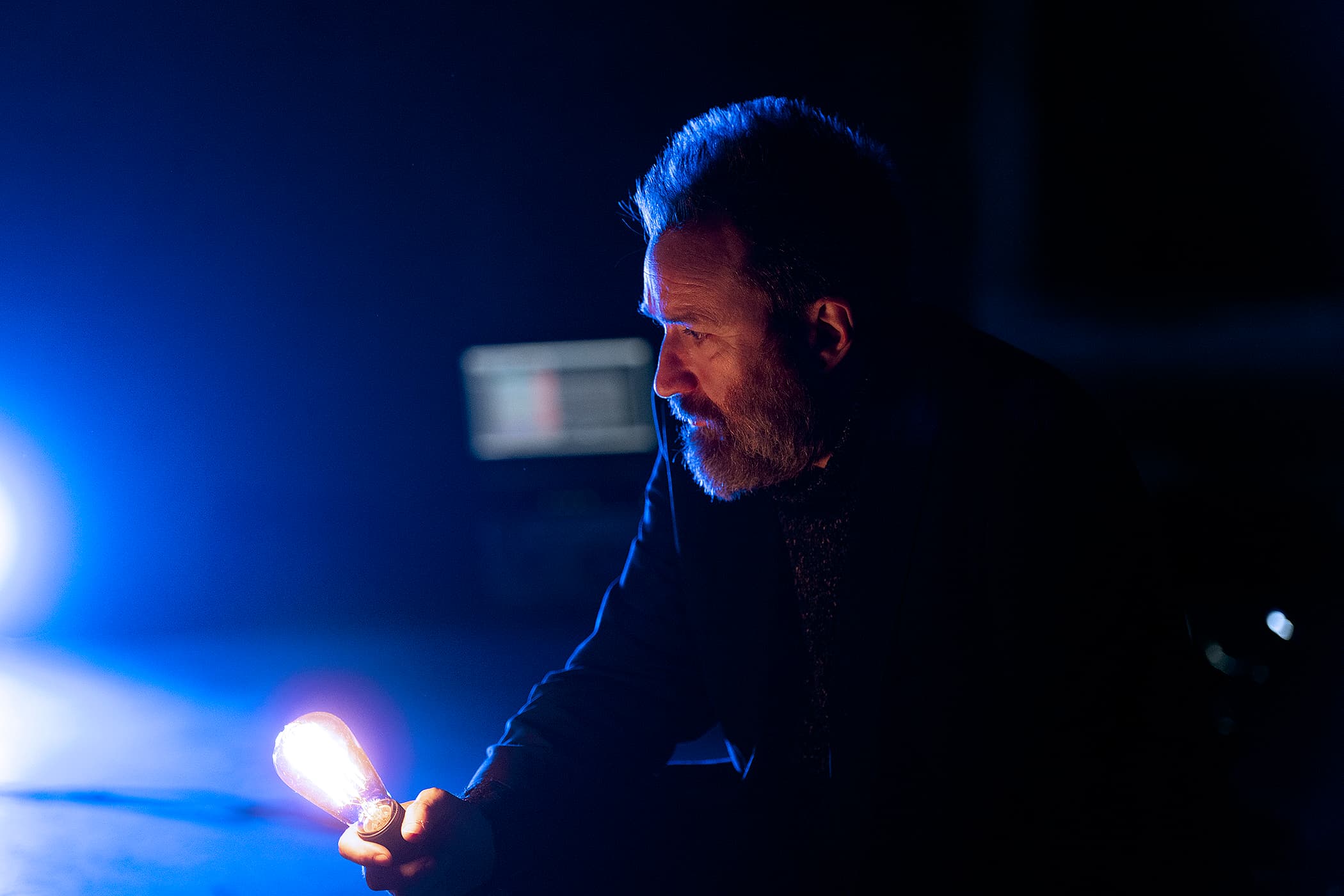 A man with a beard holds a glowing light bulb in a dimly lit environment with a blue hue.