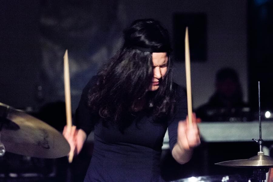 A person with long hair is intensely playing the drums in a dimly lit setting.