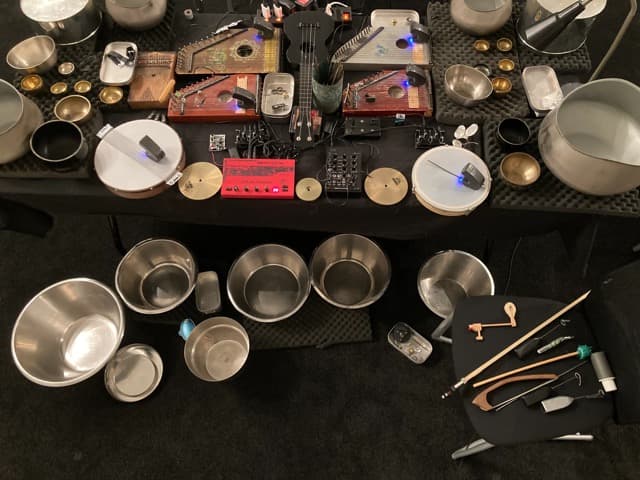 A collection of musical instruments and bowls, including string instruments, percussion items, and various metal bowls, arranged on a table.