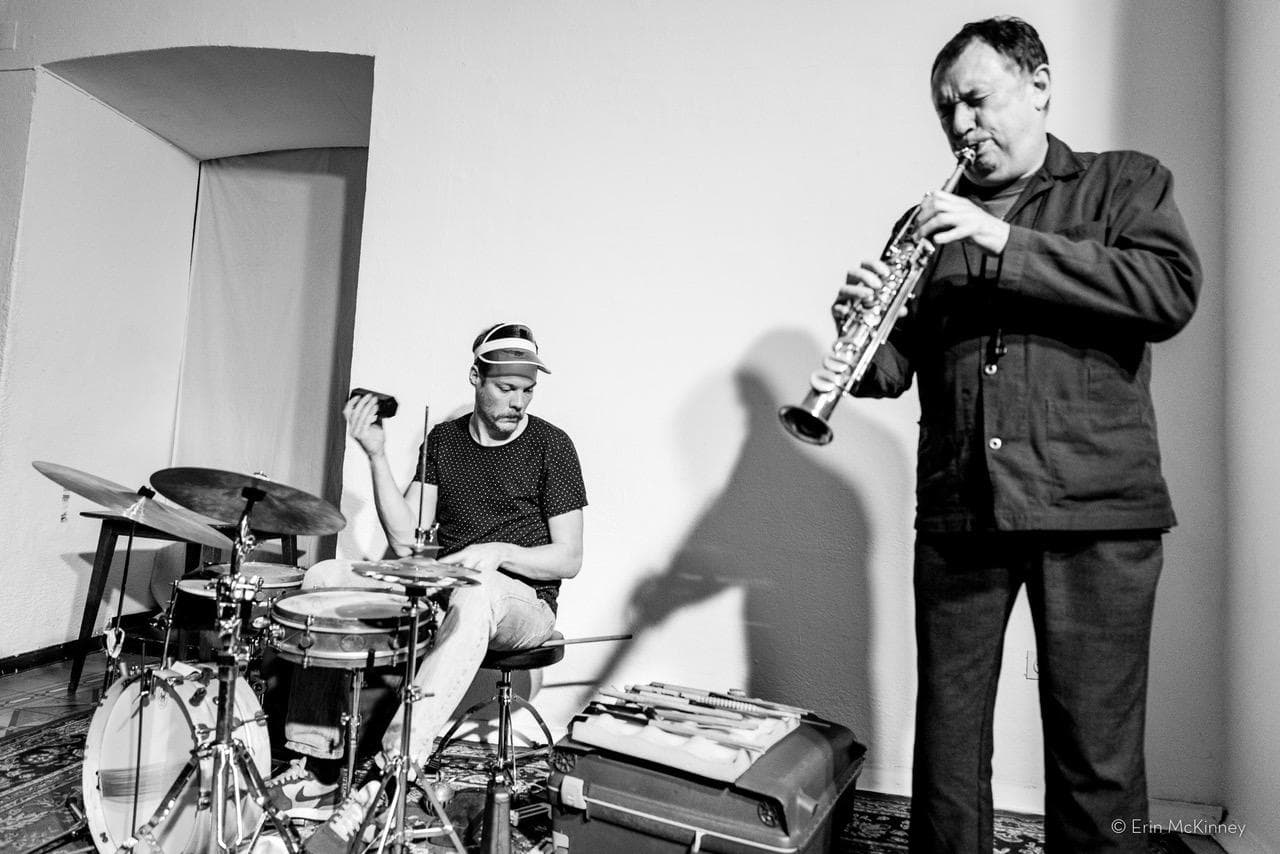A drummer plays while a saxophonist performs in a minimalist indoor setting.