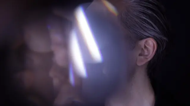 A partially obscured person with slicked-back gray hair is behind a reflective, prism-like surface, creating a distorted visual effect.