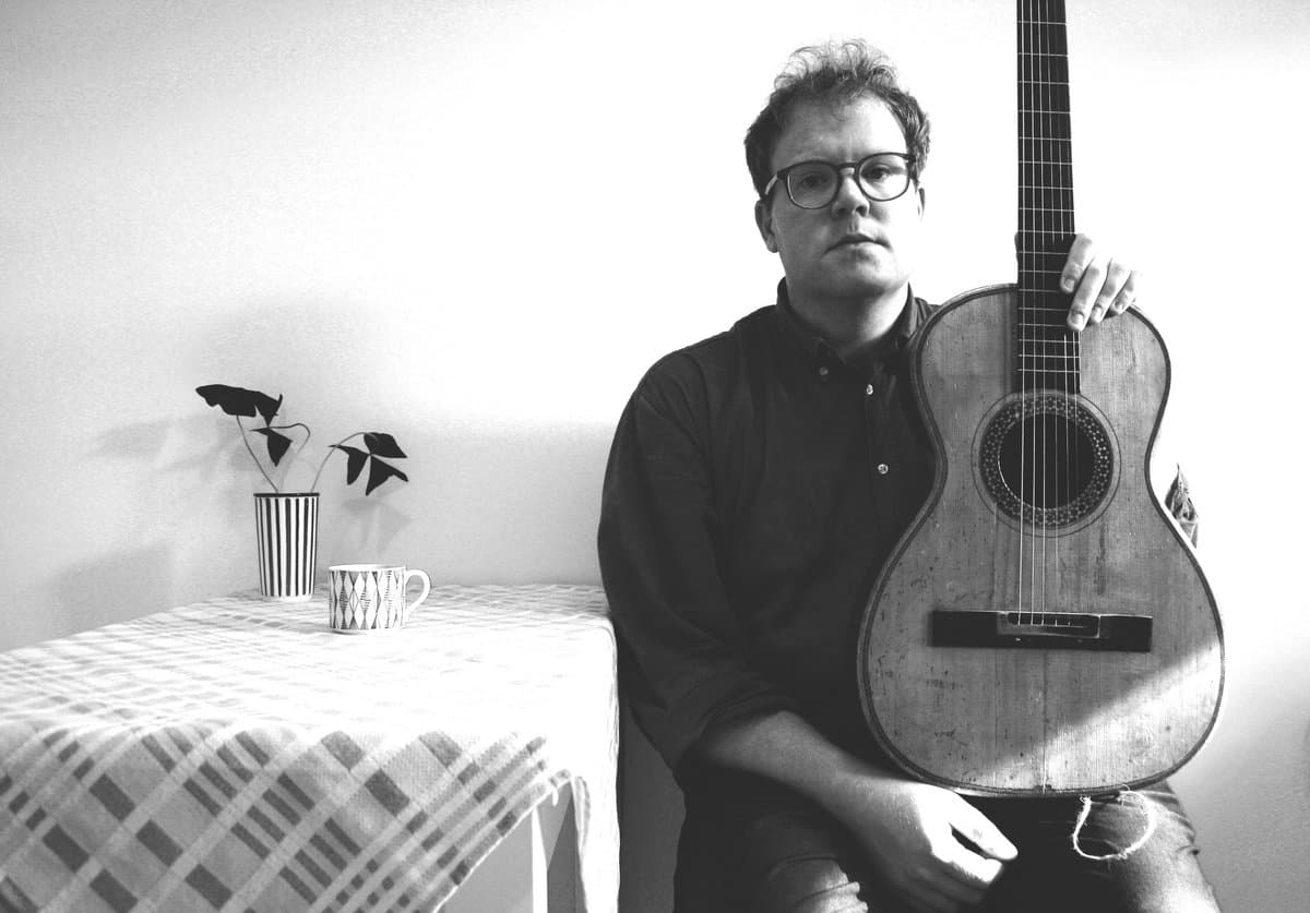 A person with glasses sits holding a guitar next to a table with a patterned cloth, a striped vase with leaves, and a geometric patterned mug.