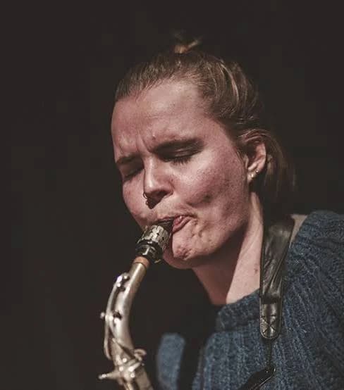 A person is playing a saxophone with a focused expression, wearing a blue sweater.