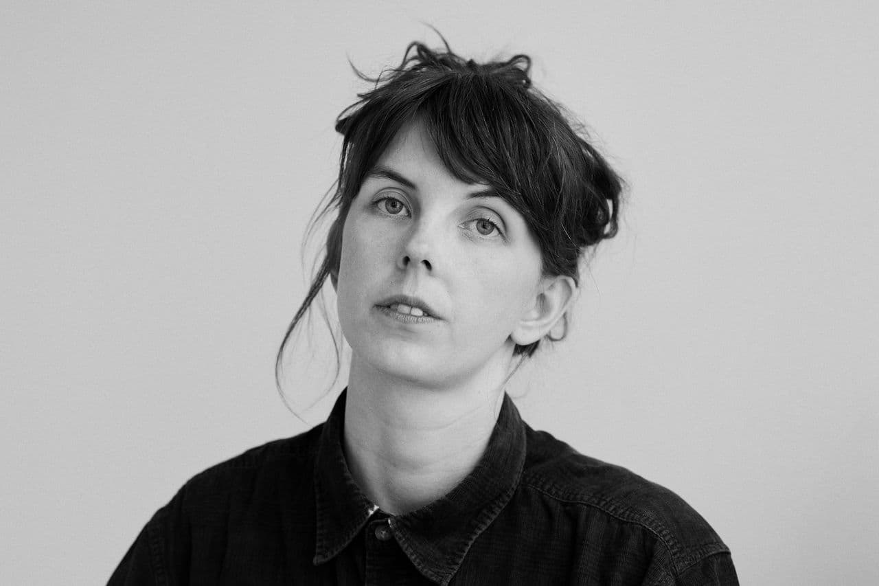A person with tousled hair and a neutral expression is wearing a dark collared shirt against a plain background.