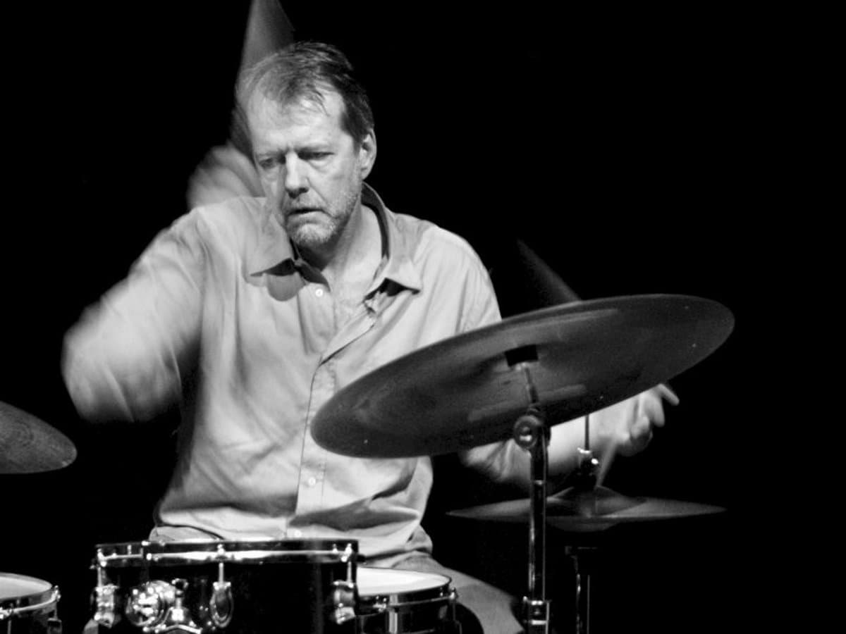 A man is playing a drum set with focused intensity in a black and white setting.