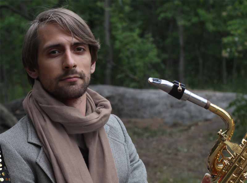 A person wearing a scarf and holding a saxophone stands outdoors with trees in the background.