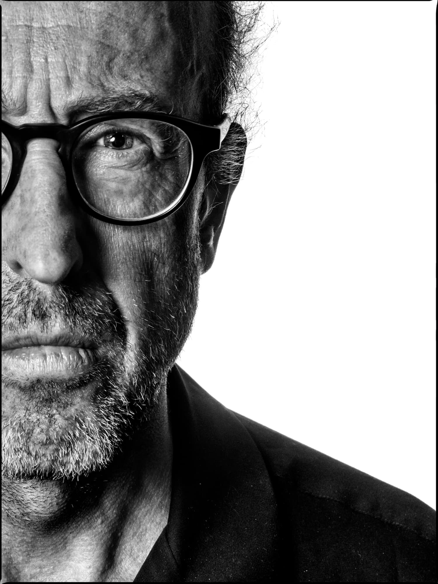 A black and white close-up of the left side of an older man's face with glasses and a beard, against a white background.