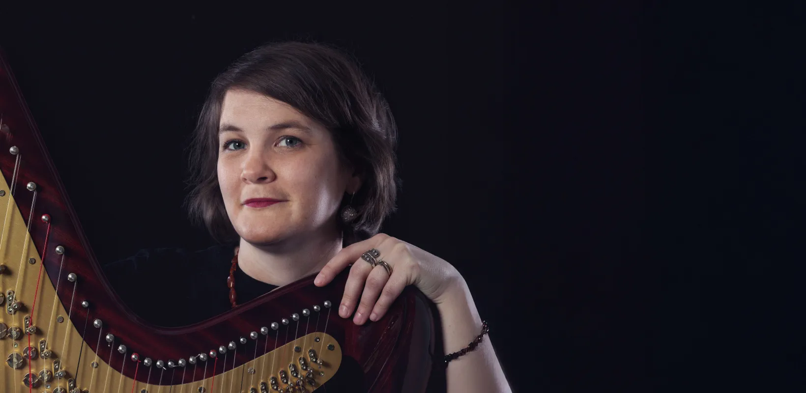 A person with short hair holds a harp against a dark background.