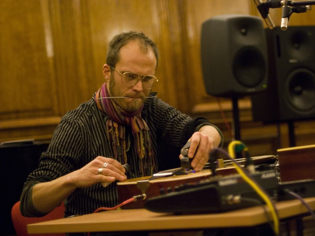 A person is playing a musical instrument with a bow, wearing glasses and a scarf, in a room with wooden walls and audio equipment.