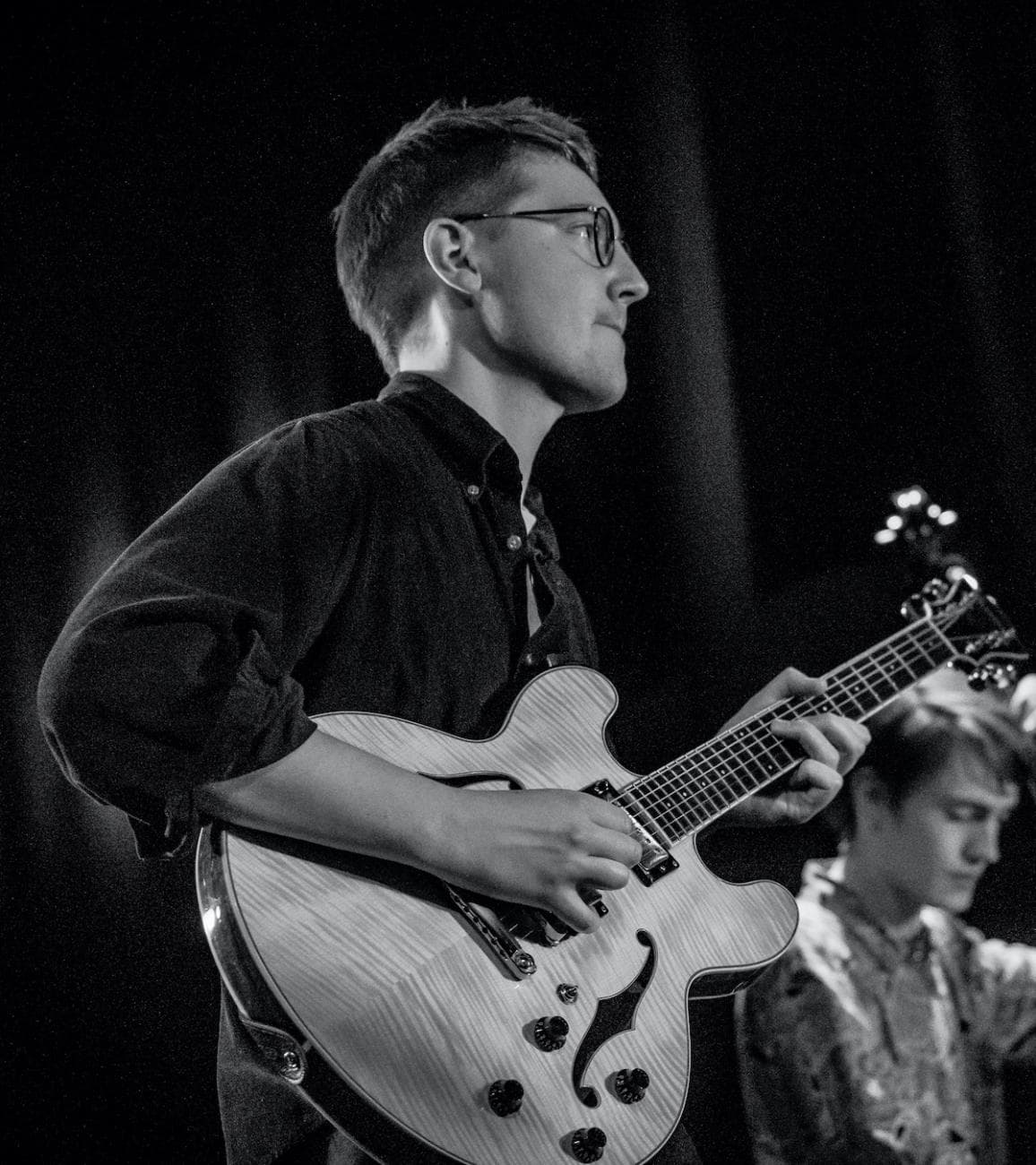 A black and white photo of a person playing an electric guitar on stage, with another person in the background.
