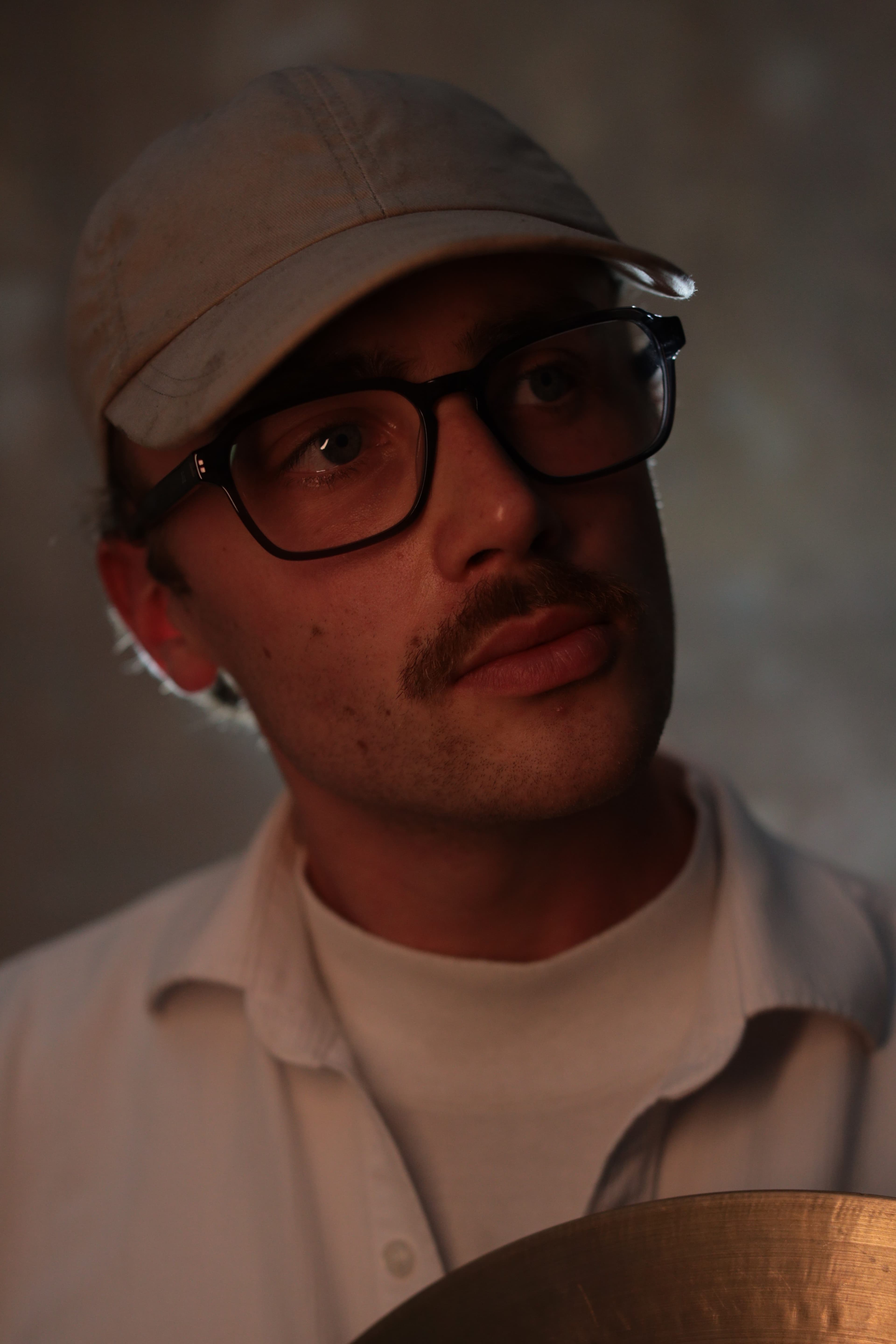 A person with glasses, a mustache, and a cap looks to the side, wearing a white shirt.