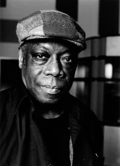 An older man wearing a cap and dark shirt is looking at the camera.
