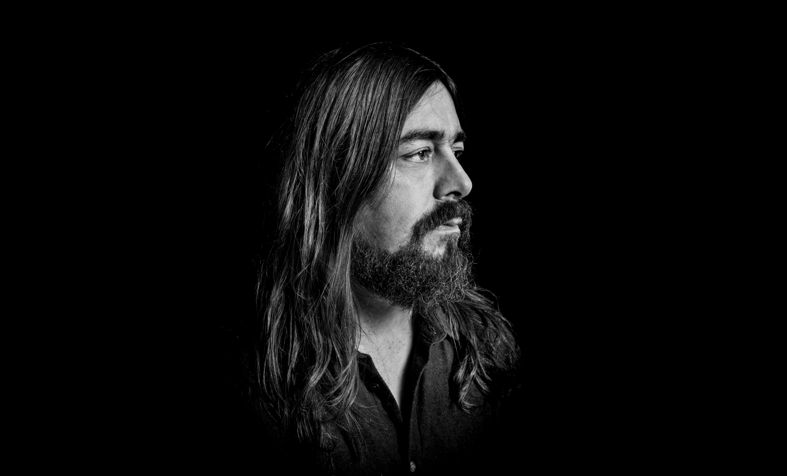 A black and white portrait of a man with long hair and a beard, gazing to the right.