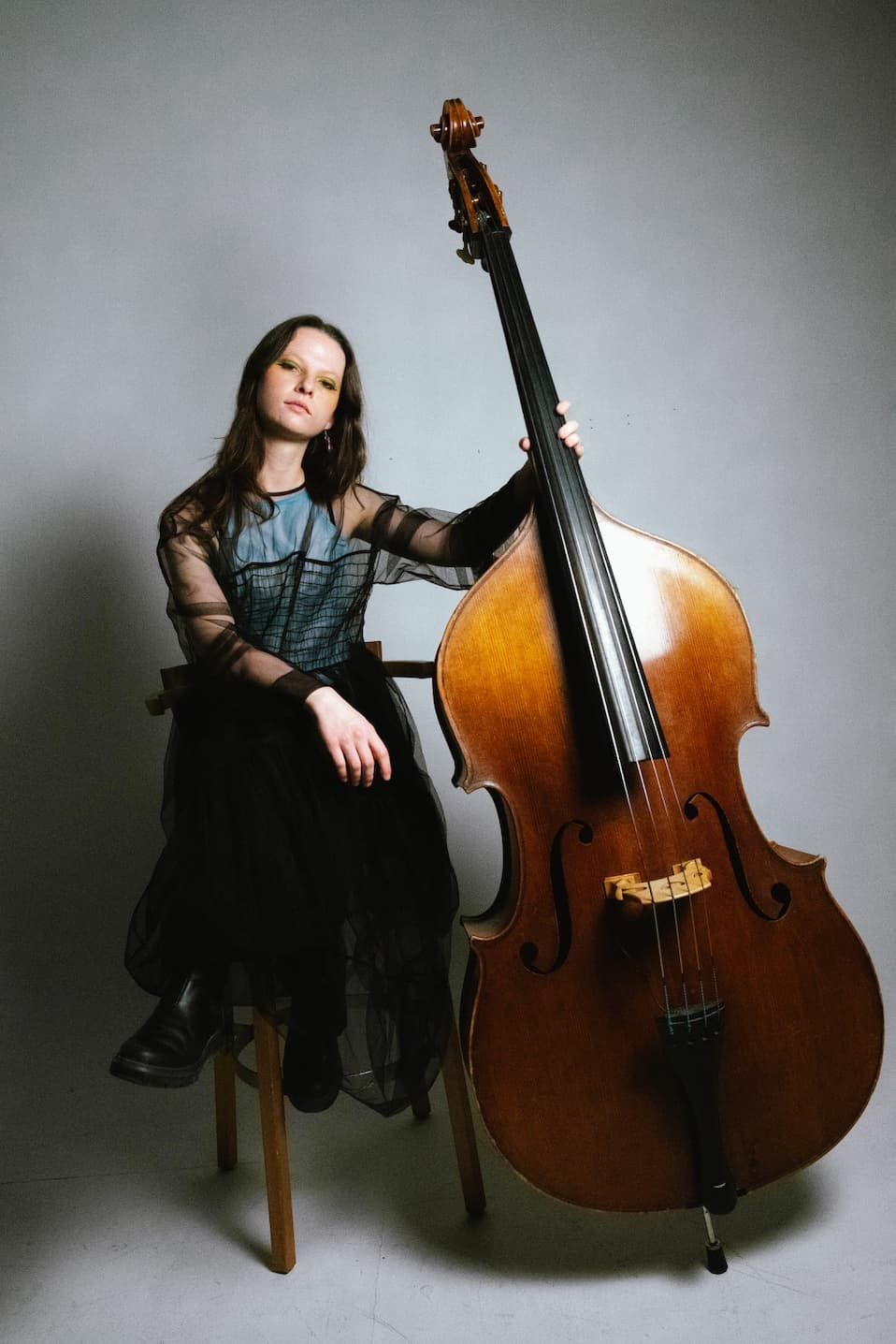 A person in a sheer black dress sits on a chair, holding a double bass upright.