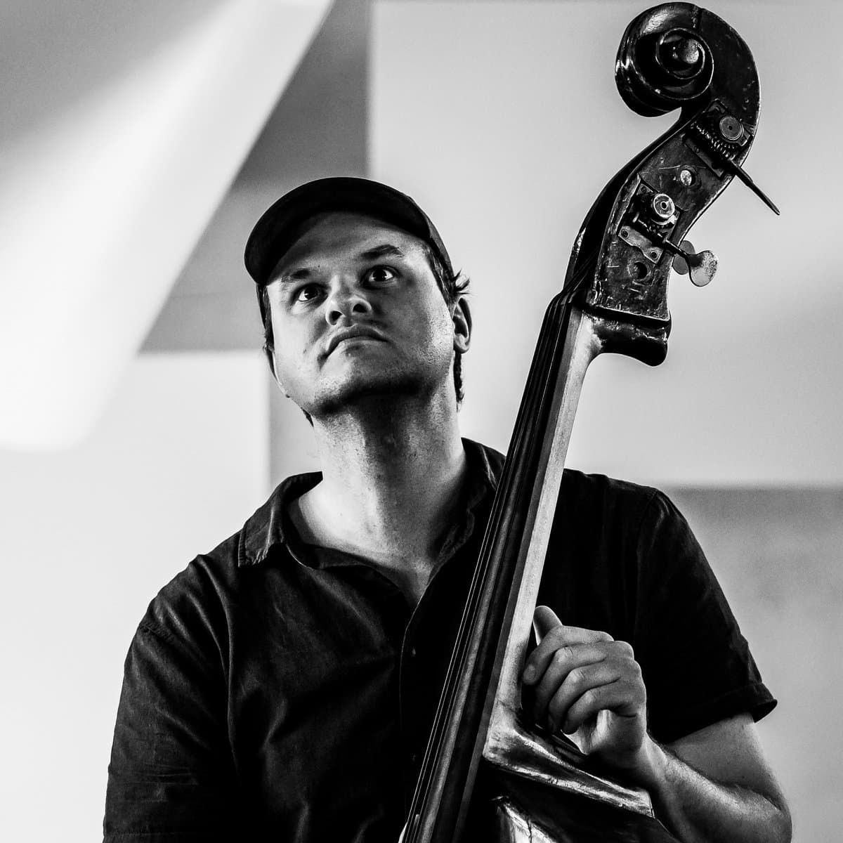 A person wearing a cap is playing a double bass, looking upwards in a black and white setting.
