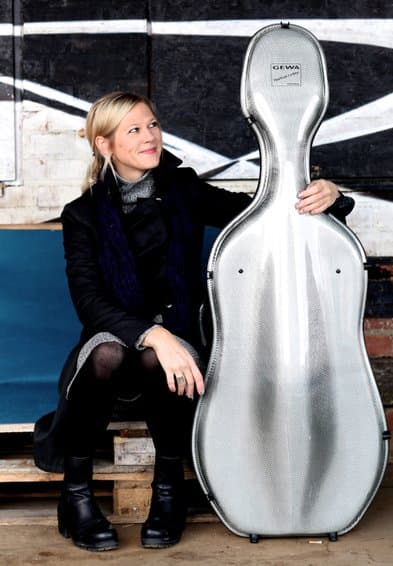A person in a coat and boots sits on a bench, holding a silver cello case.