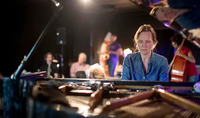 A person is playing a grand piano in a dimly lit room with other musicians and people in the background.
