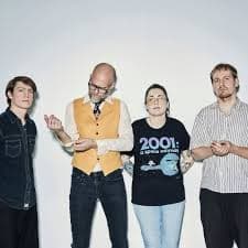 Four people stand against a plain background, each with a unique style, with one wearing a "2001: A Space Odyssey" shirt.