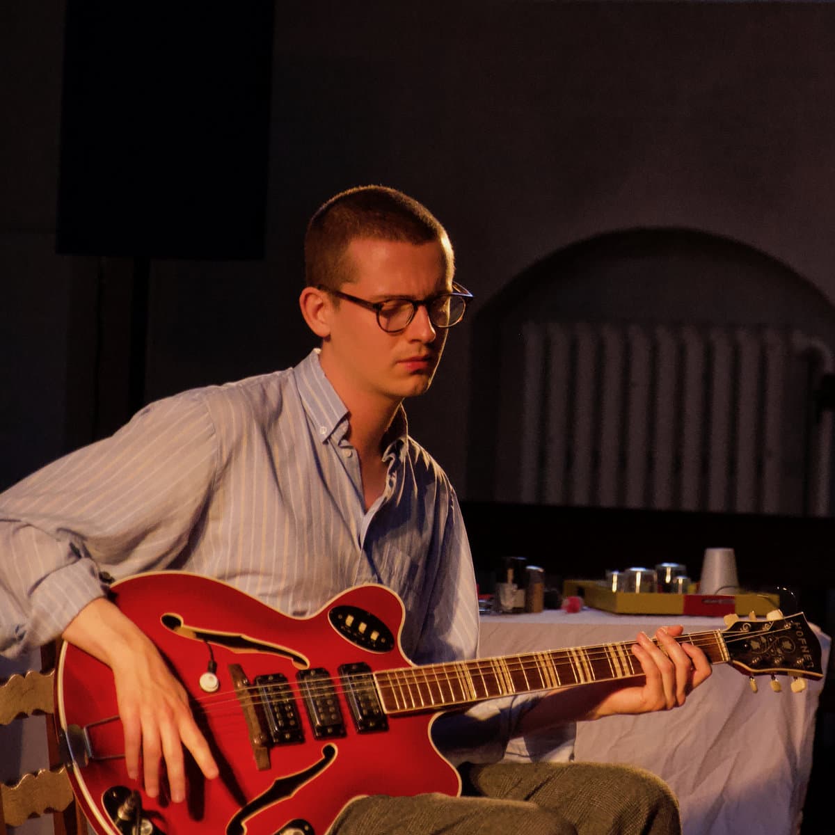 A person wearing glasses and a striped shirt is playing a red electric guitar.