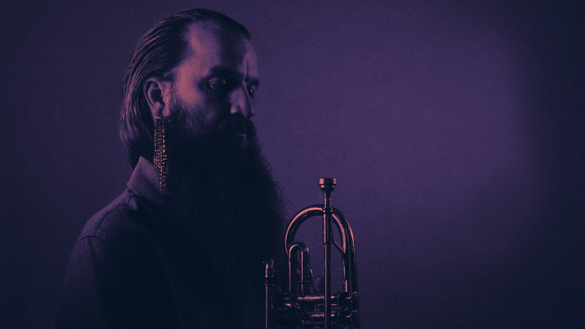 A man with a long beard and a decorative earring holds a trumpet, set against a dark, moody background with purple lighting.