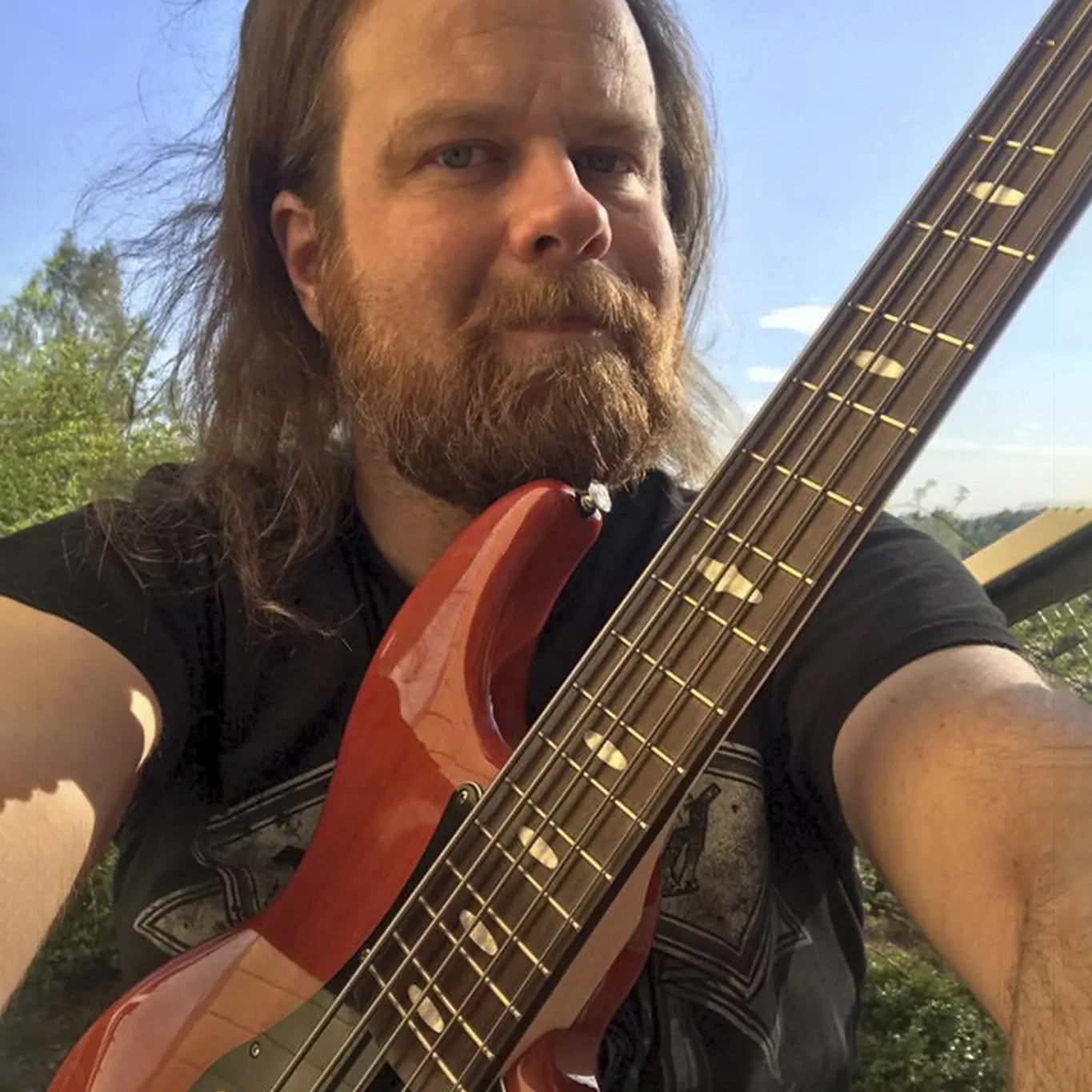 A person with long hair and a beard is holding a red electric guitar outdoors.