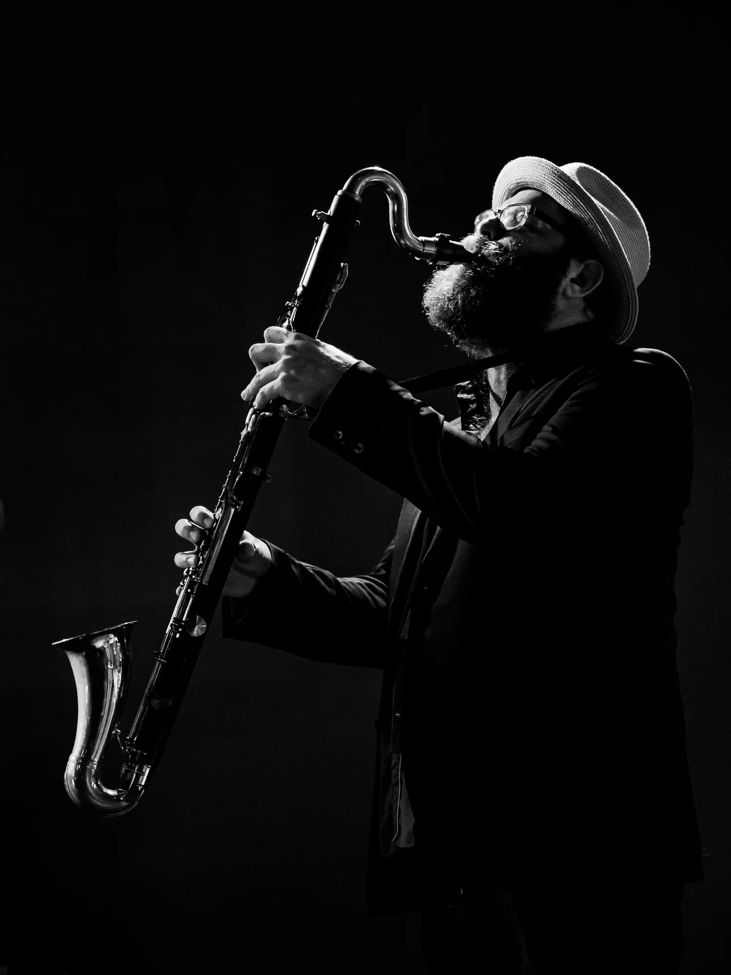 A musician in a hat and glasses plays a bass clarinet on stage, captured in a dramatic black-and-white setting.