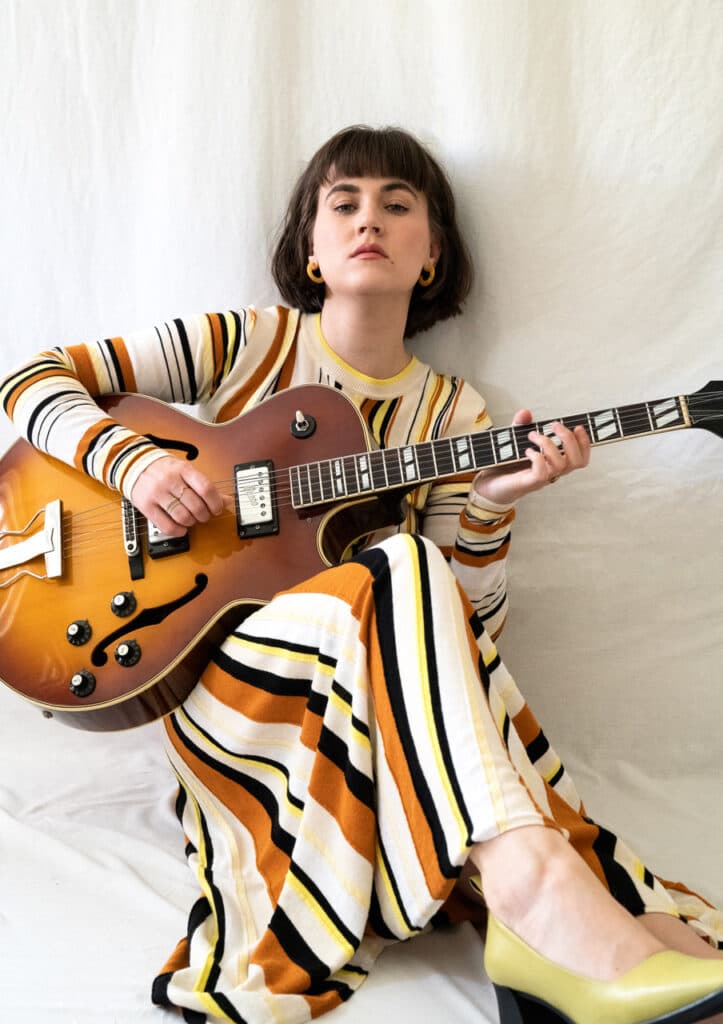 A person with short hair is sitting against a white background, holding a guitar, and wearing a striped dress with yellow, black, and orange tones.