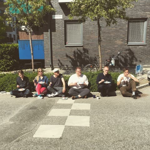 Six people are sitting on the sidewalk eating food on plates, with a brick building and bicycles in the background.
