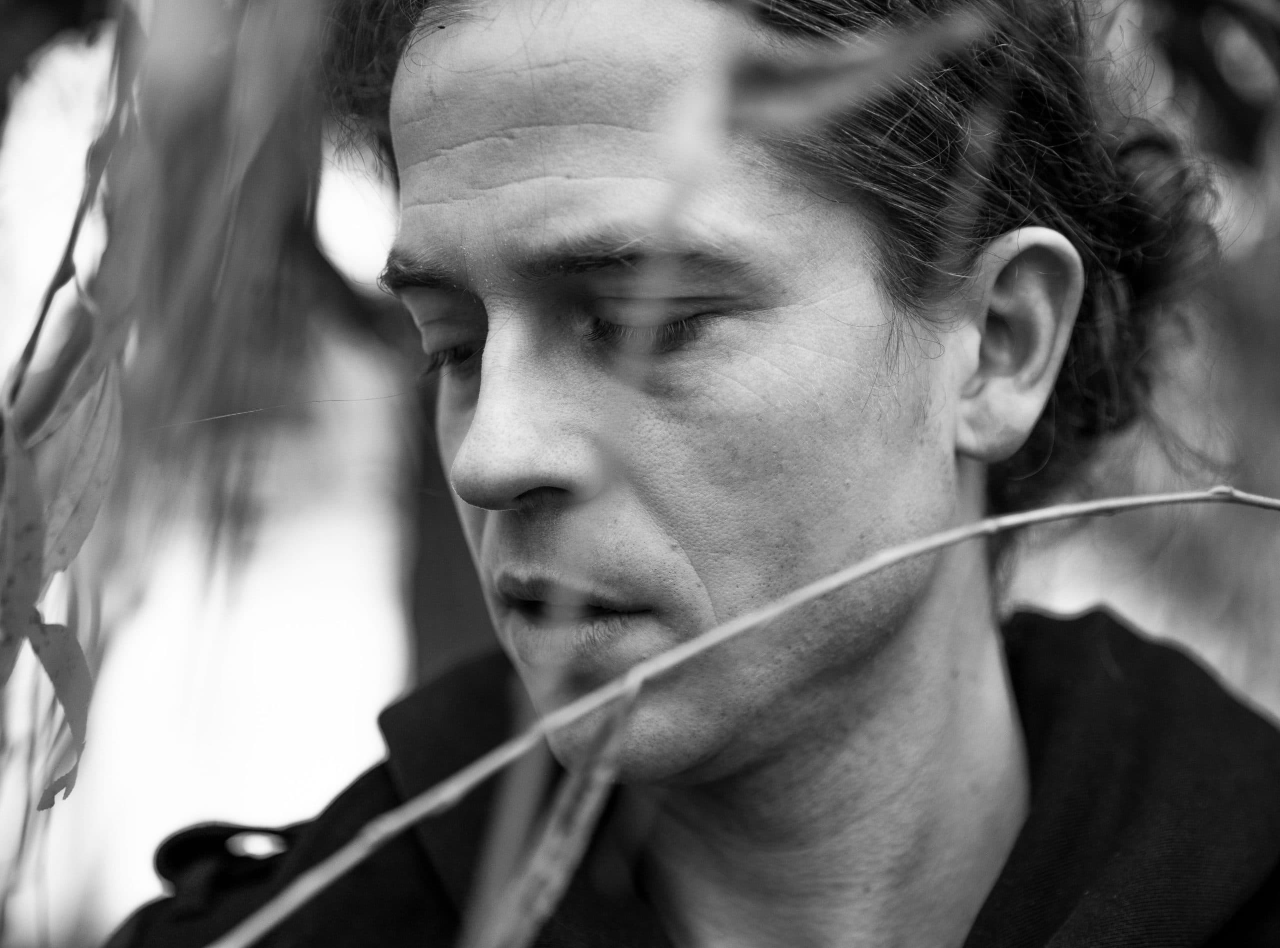 A black and white close-up of a person looking downward, partially obscured by foliage in the foreground.