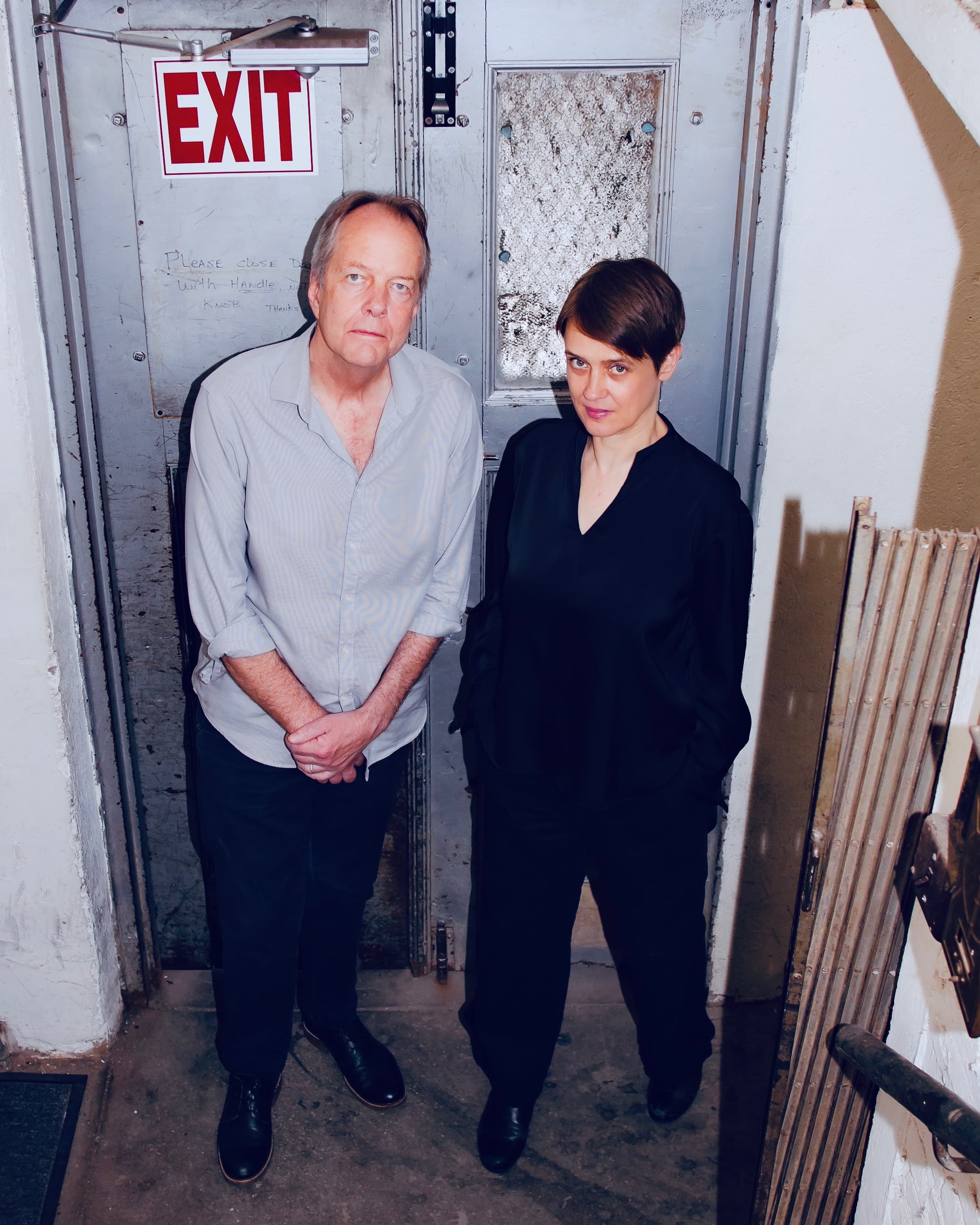 Two people stand in front of a metal door with an "EXIT" sign above it.