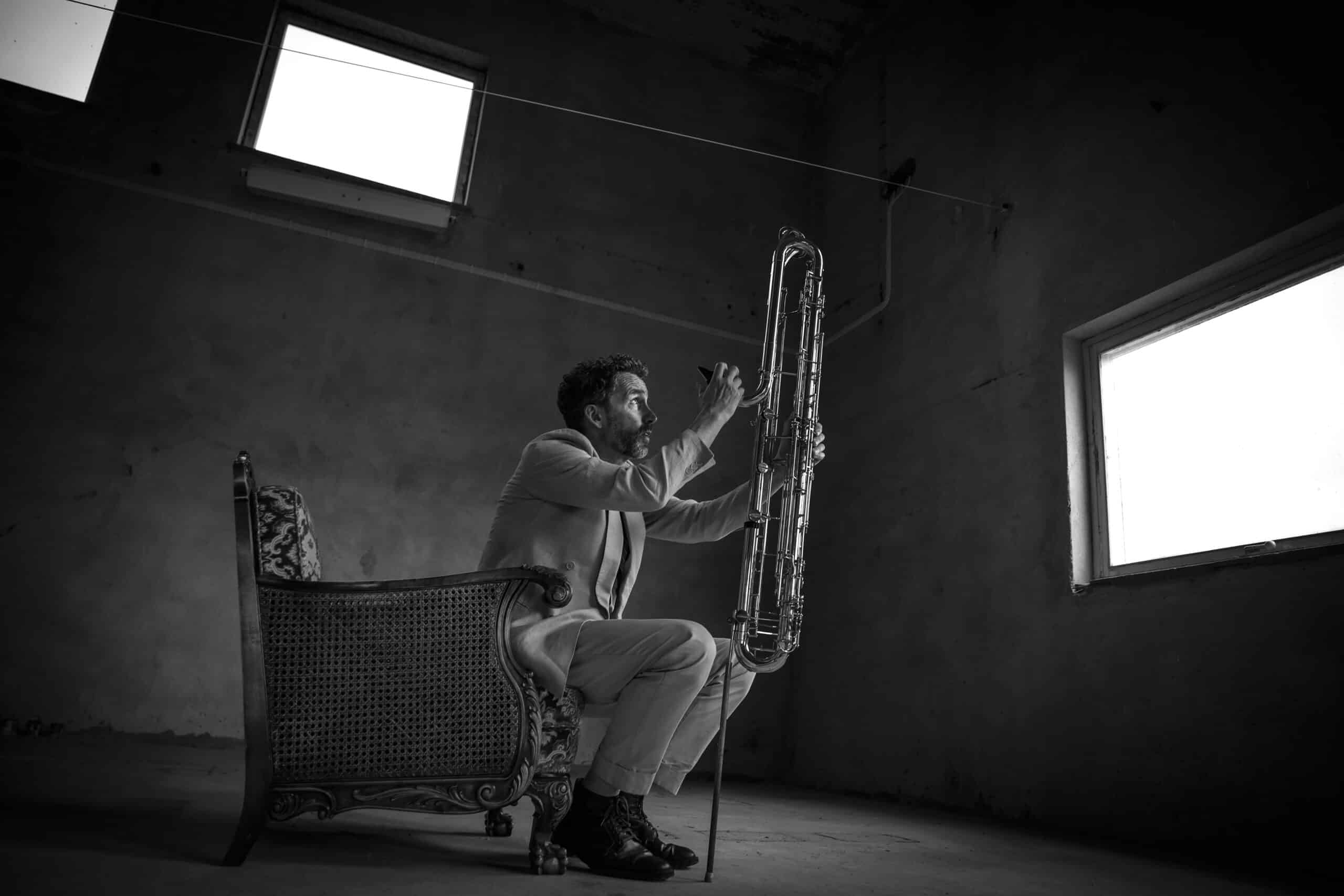 A man in a suit sits in a chair playing a large wind instrument in a dimly lit room with windows.