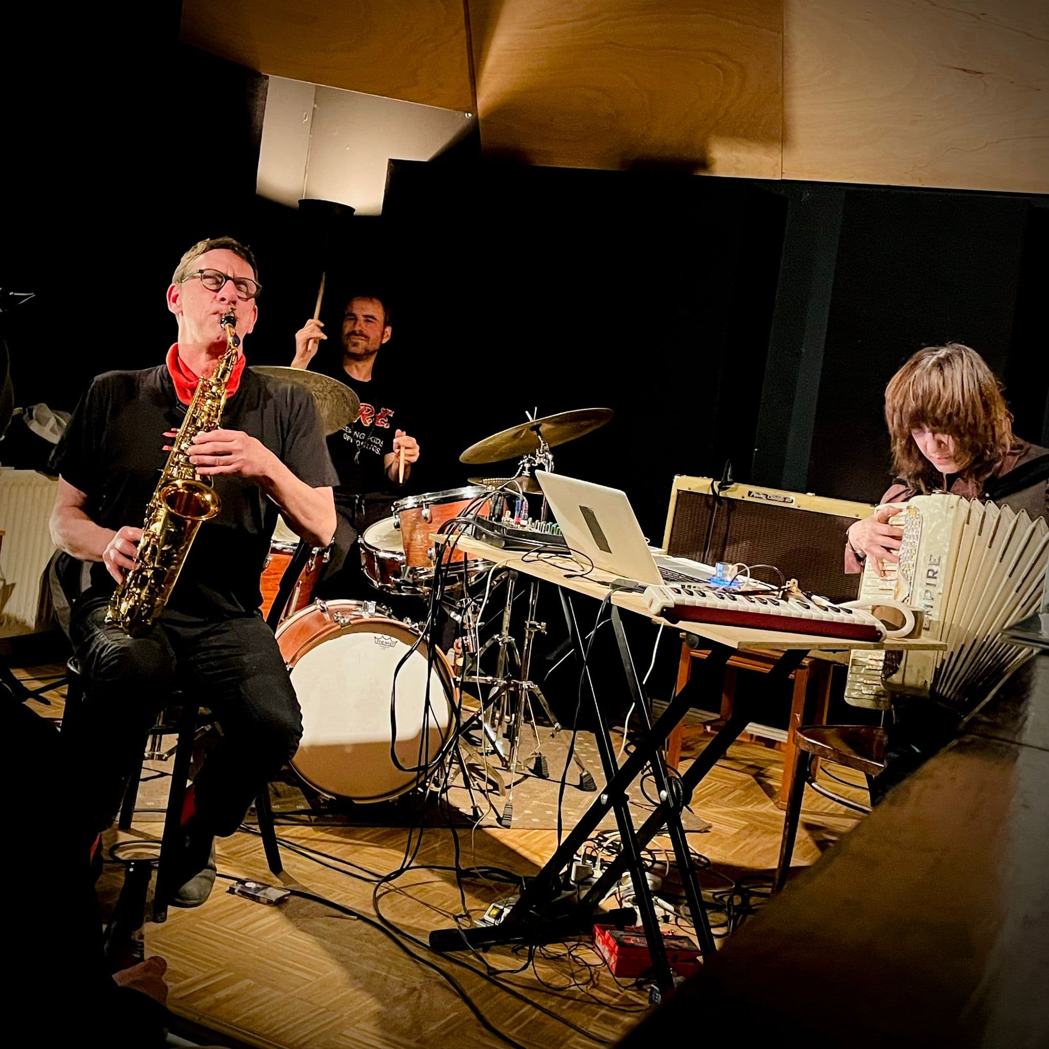 Three musicians are performing; one plays the saxophone, another is on drums, and the third plays an accordion beside electronic equipment.