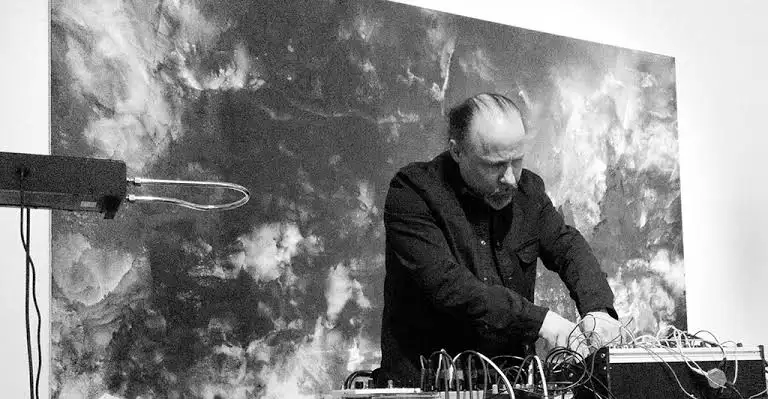 A person is adjusting electronic music equipment in front of a cloudy abstract backdrop.