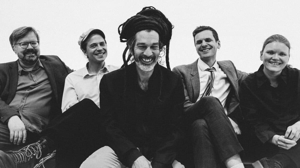 Five people are sitting and smiling, with the central figure wearing distinctive long dreadlocks.