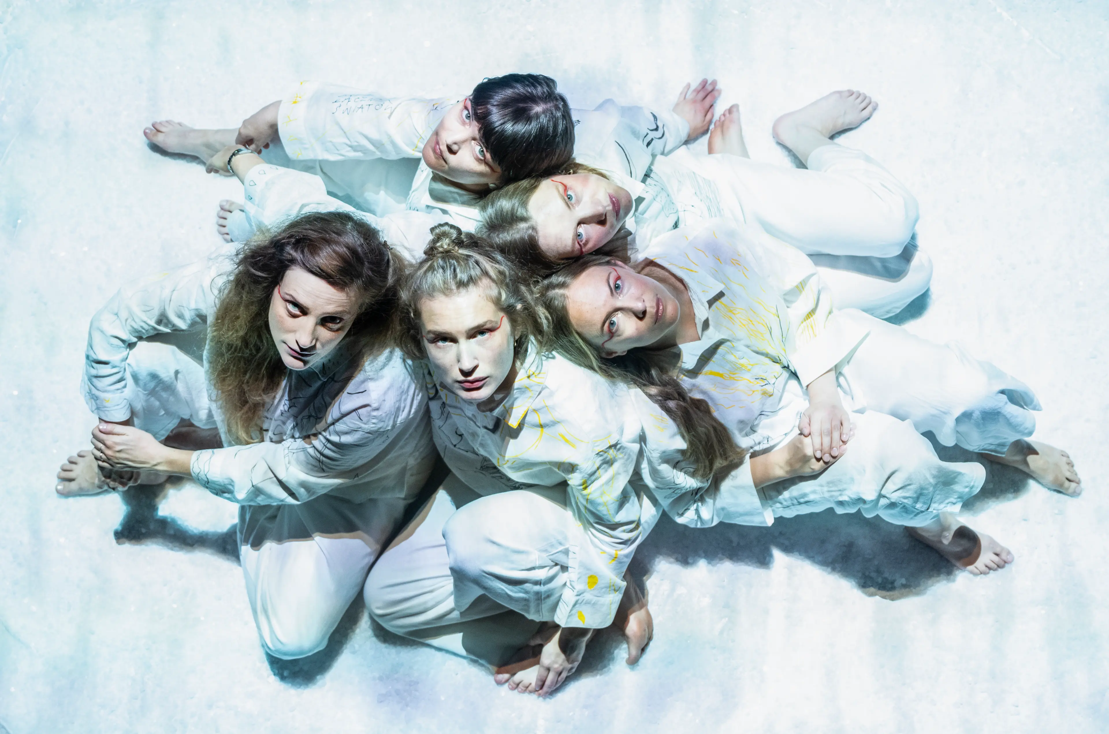 Five people dressed in white lie on a white surface, looking up towards the camera.