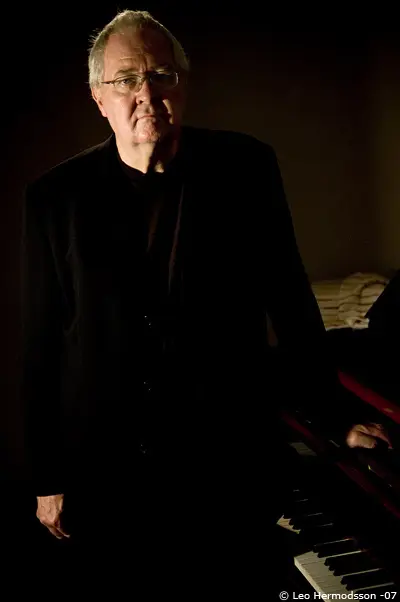 An older man in a dark suit stands next to a piano, with shadows cast in a dimly lit room.