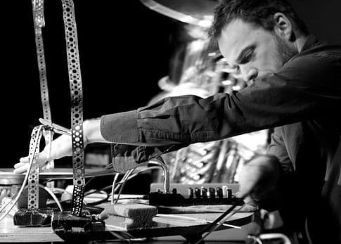 A person is intensely focused on manipulating audio equipment and wires on a table, with a musical instrument visible in the background.