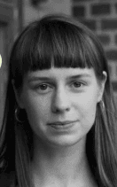 A person with straight hair and bangs is looking at the camera, wearing hoop earrings.