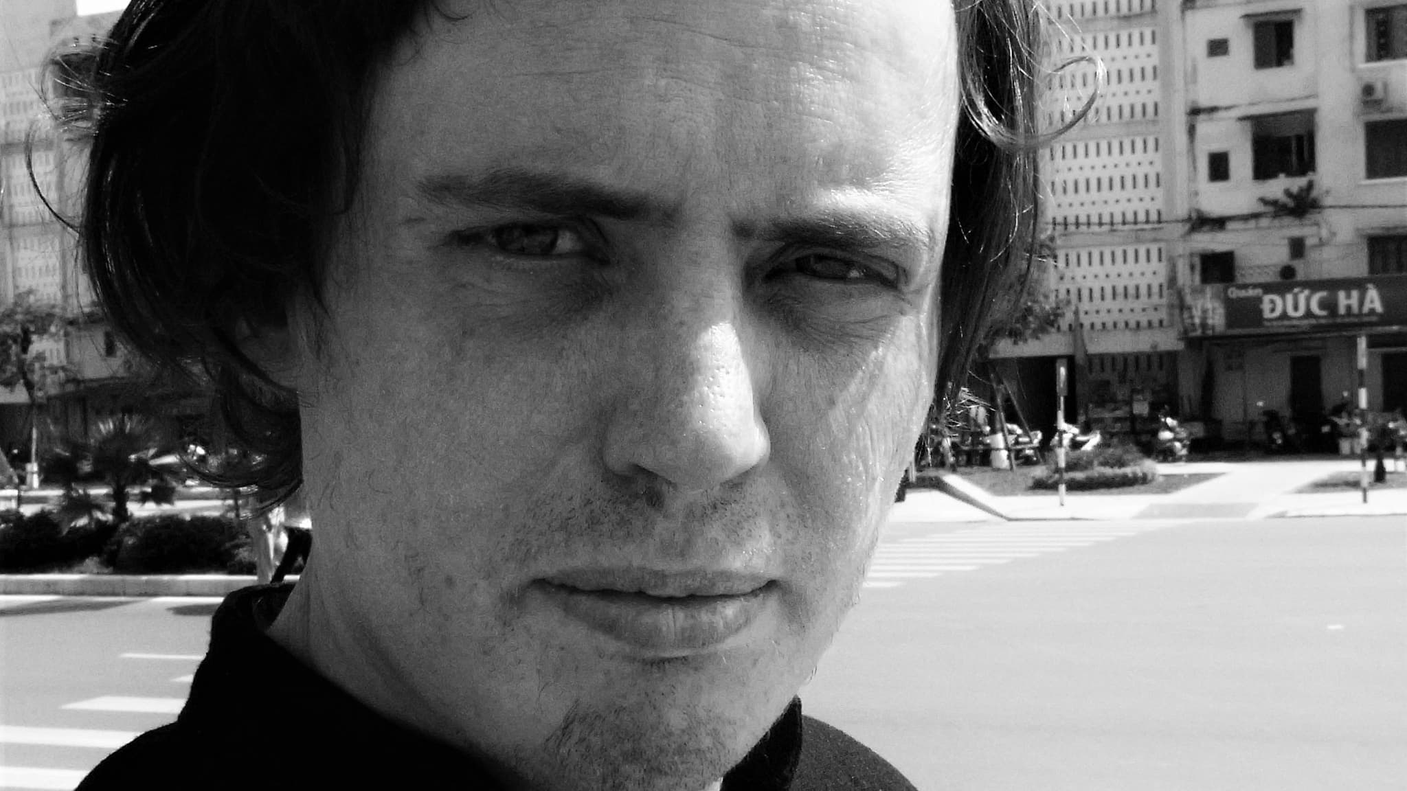 A person with medium-length hair is looking into the camera, standing outdoors near a street with buildings in the background.