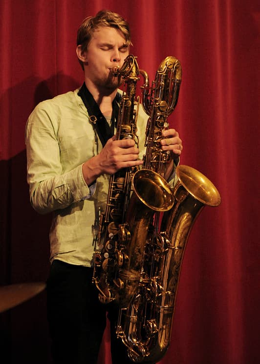 A person is playing two saxophones simultaneously on a stage with a red curtain backdrop.